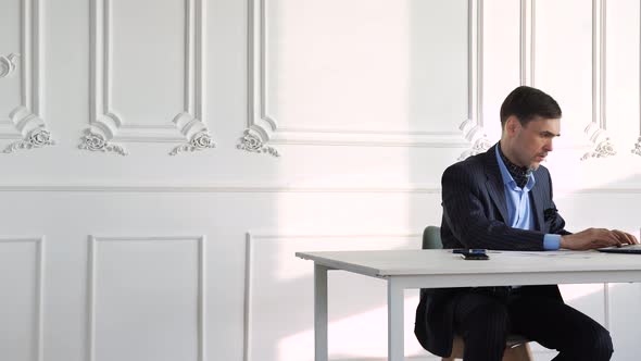 Man Works at a Laptop in a Suit Sitting at a Table in a Large Hall alt