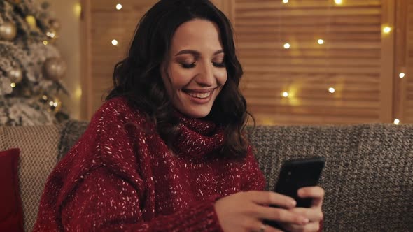 Attractive Woman Using Smartphone Sitting on Couch, Messaging, Smiling in Decorated Apartment Near alt