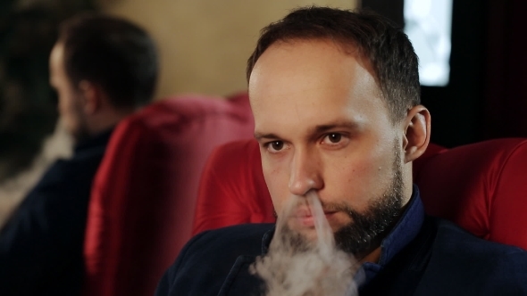 Brutal Guy With a Beard Smokes An Electronic Cigarette In a Red Armchair alt
