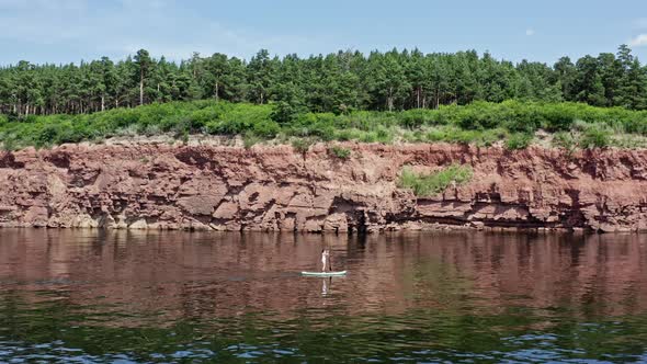 Drone Shot of a Woman Floating on a Board with a Paddle Along a Rocky Cliff
