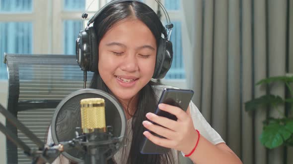Asian Kid Girl Holding Phone And Wearing Headphones Singing While Recording Podcast In Studio alt