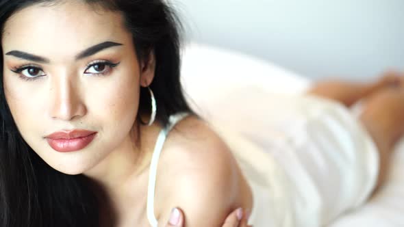 Beautiful and Sexy Young Asian Woman Posing to Camera in White Dress in Bed alt
