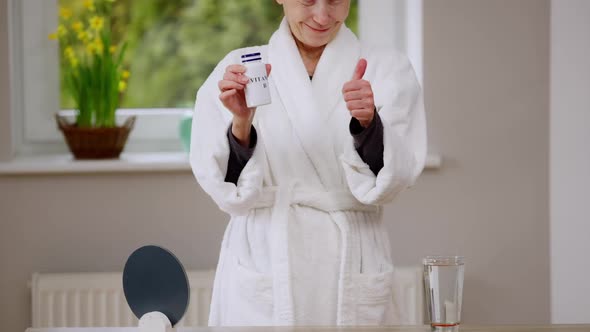 Unrecognizable Slim Mature Woman in White Bathrobe Gesturing Thumb Up Smiling Showing Bottle with alt