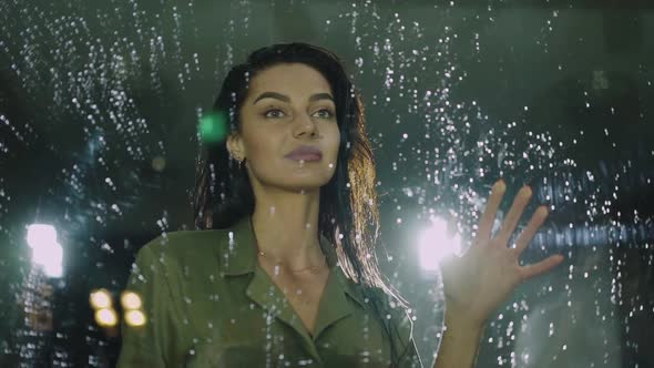 Beautiful Woman Touches the Glass Door with Rain Drops and Smiling alt