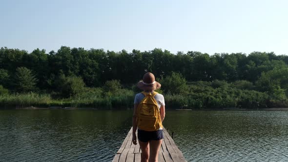 Young Tourist Woman in Hat and with a Backpack is on a Wooden Bridge Over the River alt