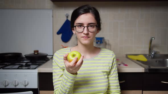 The girl in glasses sits at the kitchen table and eats an apple, looks at the camera alt