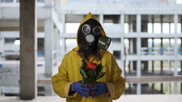 Woman in Gas Mask and Yellow Overalls Found Flower Life Was Born Shows and Holds It Out to Camera alt