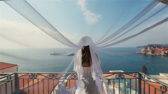 Charming Blond Bride In Long Bridal Veil Standing On Terrace With The Sveti Stefan In Montenegro