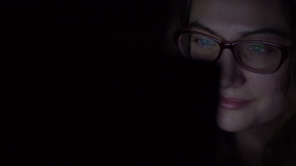 Young Girl in Glasses Uses a Laptop While Lying on the Bed. Night Shooting alt