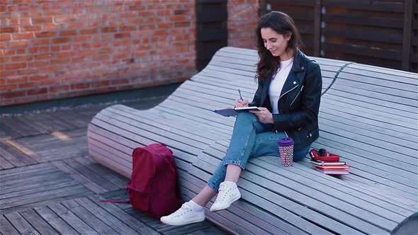 Female Student Writing in Notebook Sitting Outside on the Modern Wooden Bench alt