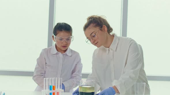 Girl With a Teacher in Lab Coats and Safety Glasses Conducts Experiments alt