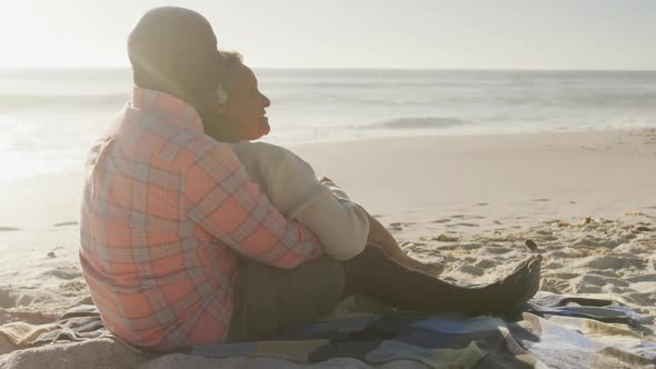 Smiling senior african american couple embracing and sitting on sunny beach alt