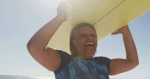 Happy senior african american woman walking with surfboard on sunny beach alt