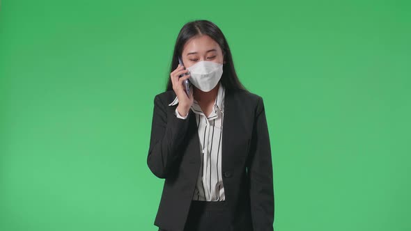 Asian Business Woman Wearing Mask Talking On Mobile Phone While Walking On Green Screen Studio alt