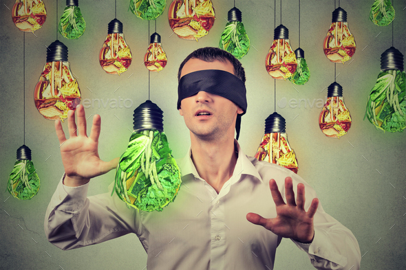 Blindfolded man walking through light bulbs shaped as junk food green vegetables