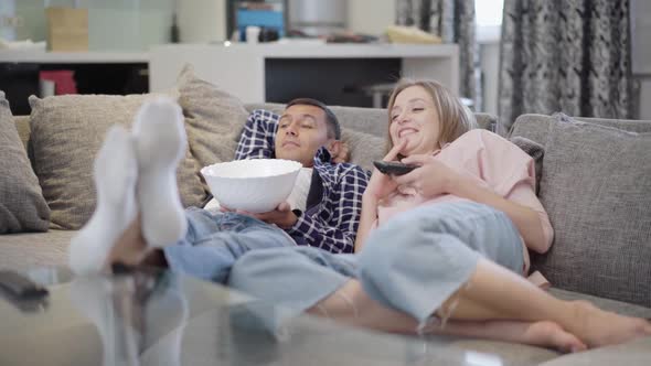 Wide Shot of Relaxed Carefree Couple Lying on Couch Watching Comedy Movie on TV Eating Popcorn alt