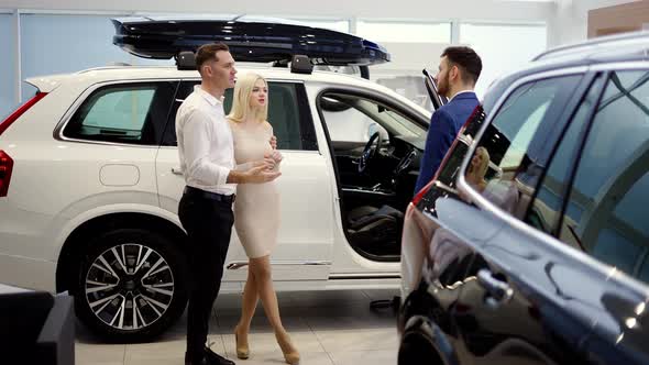 Couple Talking to Car Dealer in Dealership Discussing Automobiles Looking at Luxurious New White alt
