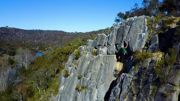 Stationary/hovering drone footage of an adult male rock climbing with a scenic creek and mountain in alt