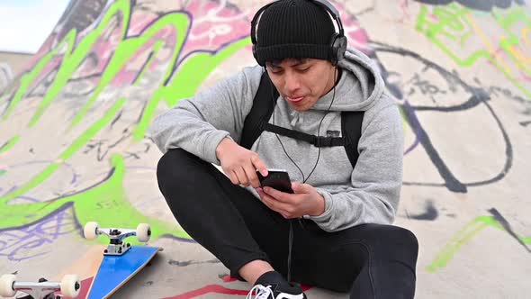 Young Latin Man Skateboarder, with Mobile Phone, Texting at Skate Park alt