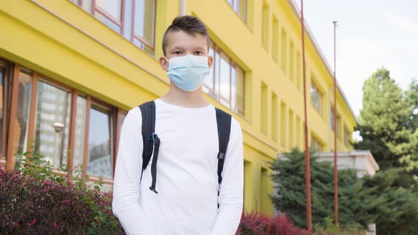 A Caucasian Teenage Boy Puts on a Face Mask and Looks at the Camera  a School in the Background