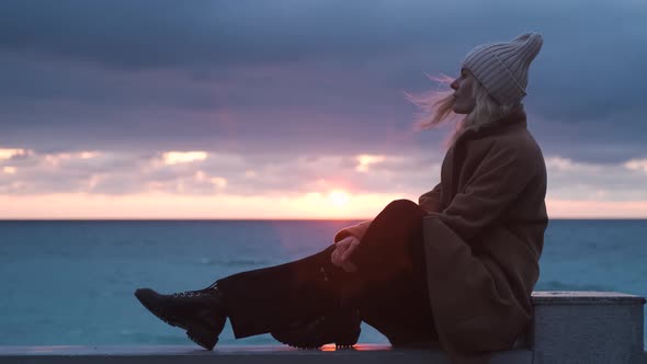 woman in a coat sitting on the beach at sunset