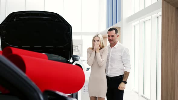 A Man Make Surprise to a Blonde Woman a New Car with Red Bow in a Showroom alt