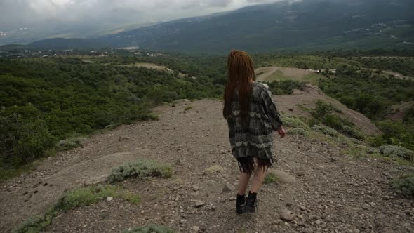 A Young Woman with Pigtails in a Light Cape Descends From the Mountain