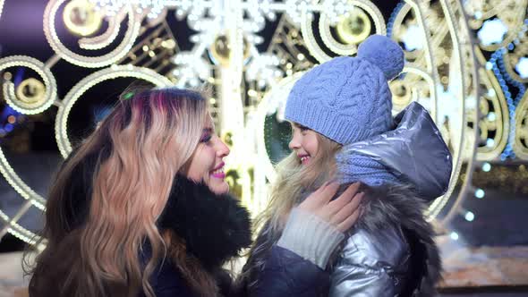 Young Mother Embracing Her Cute Daughter at Festive Street Decorations Background alt