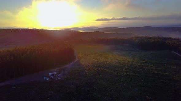 Drone footage of sunrise over a forest covered in patches of fog. People can be seen camping in bott alt