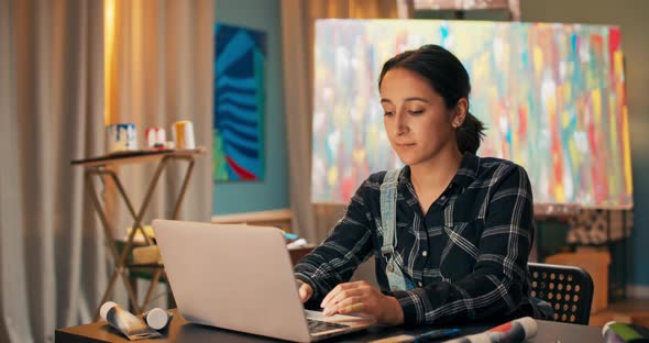 A Smiling Brunette Sits at a Desk in Front of a Laptop in Art Studio alt