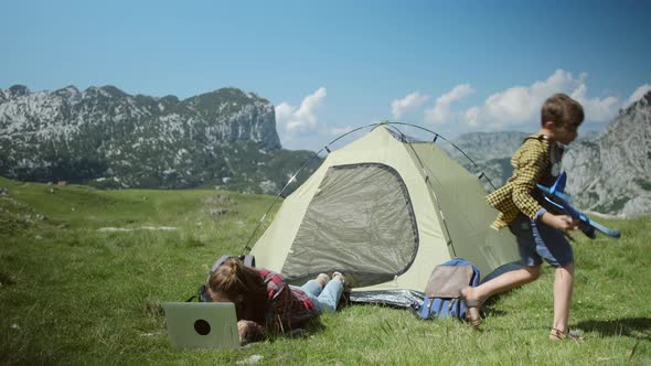 Freelancer woman working on laptop and have a rest near tent and her son playing  with a toy alt