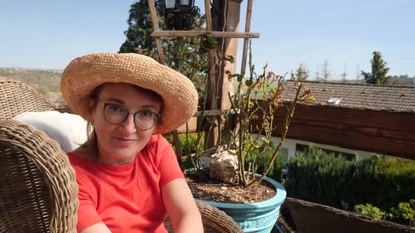 Smiling redheaded woman wearing straw hat sitting on balcony alt