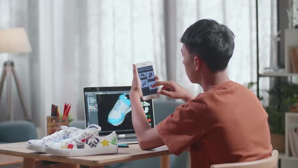 Asian Boy Footwear Designer Looking At Photos On Smartphone While Designing Shoe On A Laptop At Home alt