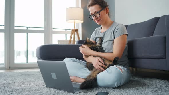 Casually Dressed Woman Sits on a Carpet with a Laptop Holds on Her Knees and Strokes a Fluffy Cat alt
