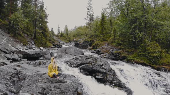 Young Woman in Yellow Raincoat Doing Yoga Near a Waterfall in the Mountains