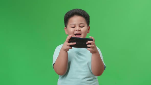 Laughing Asian Little Boy Looking At The Phone While Standing On Green Screen In The Studio alt