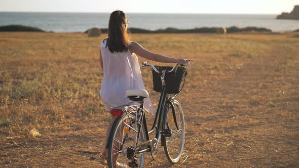 Wide Shot Happy Young Woman Walking with Bike at Sunset on Yellow Farm on Cyprus Looking Back in alt