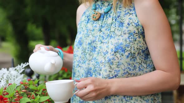 Woman in a Summer Cafe Pours Tea Into a Cup Close-up Standing. Woman Stands on the Background of the alt