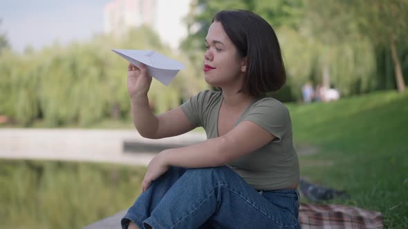 Portrait of Happy Carefree Caucasian Little Woman with Paper Plane Sitting on Lake Shore in Summer alt