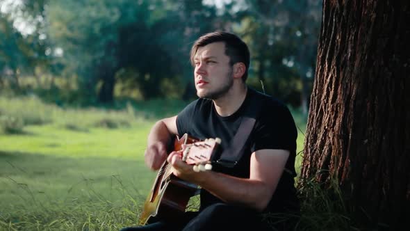 A Man in a Black Tshirt Sits on the Grass Plays and Sings a Song on an Acoustic Guitar
