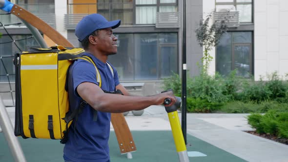 Smiling Afroamerican Man Courier Delivery Walks Street with Electric Scooter alt