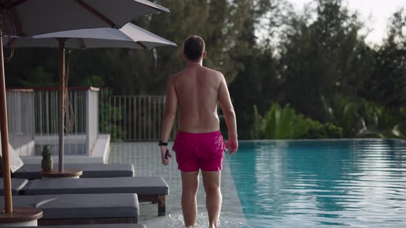 Back View of an Adult Man in Pink Swimwear Trunks Walking By the Lounge Swimming Pool at Sunset in alt