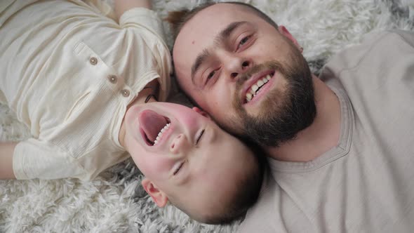 Fun daddy with beard and his little son lying at the white fur carpet and have interesting conversat