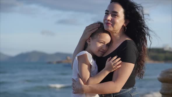 Happy Family, Mother and a Cute Daughter in Their Gentle Embrace Sit on a Bench Near the Sea, the alt