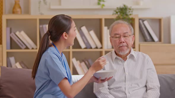 Young Asian nurse feeding breakfast to senior man at couch. Old Asian man with white beard enjoy his alt