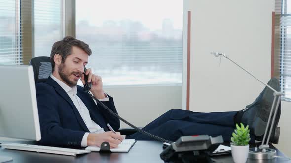 Young Businessman Sitting at the Workplace in Office and Talking on the Phone with His Legs on the alt