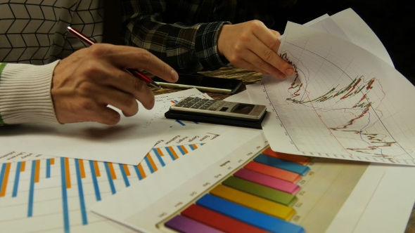 Young Businessman And Businesswoman Analyzing Graphs, Making Calculations And Taking Notes alt