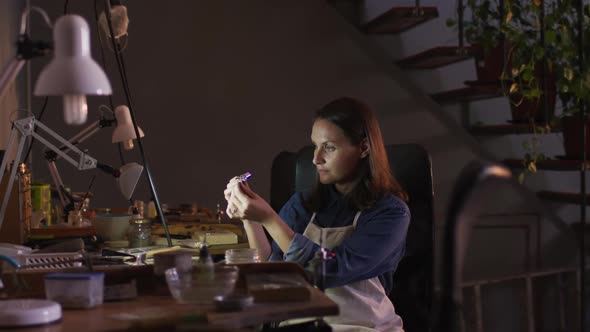 Profile of focused caucasian female jeweller sitting at desk, making jewelry in workshop alt