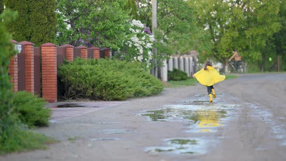 Little Sweet Girl in Yellow Raincoat and Boots has fun and Runs through the Puddles on the Road.