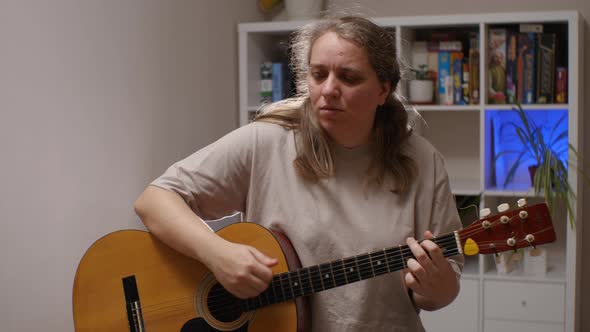 A woman with a serious face plays an acoustic guitar sitting in a room alt